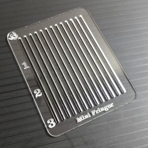 Puede incluir: Una herramienta de plástico transparente con una cuadrícula de líneas y los números 1, 2 y 3. El texto "Mini Fringer" está impreso en la herramienta.