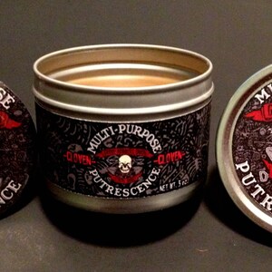 May include: A silver tin with a black and white label that reads "Multi-Purpose Grave Robbers Union Local 666 Putrescence" and features a skull and crossbones with shovels. The tin is open and shows a brown substance inside.