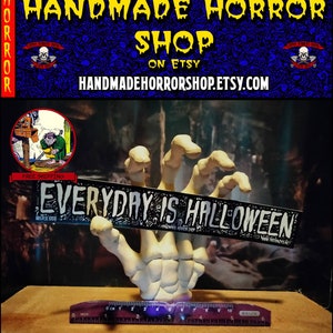 May include: A white skeletal hand holding a black sticker that reads "EVERYDAY IS HALLOWEEN". The hand is on a wooden surface with a ruler for scale. The sticker is decorated with a skull and crossbones pattern.