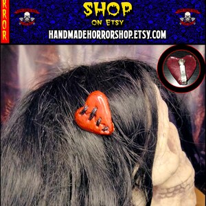 May include: A red heart-shaped hair clip with black stitching and a distressed look. The clip is made of polymer clay and is handmade. The clip is shown in a close-up image on a person's hair.
