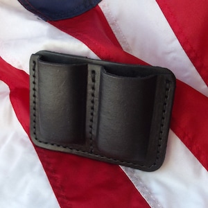 May include: A black leather double magazine pouch with stitched seams. The pouch is designed to hold two magazines.