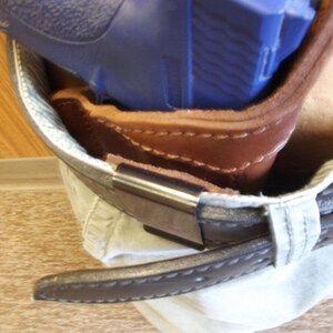 May include: A brown leather holster with a blue handgun inside, attached to a belt. The holster is stitched with visible seams and has a metal clip. The belt is dark brown with a stitched edge.