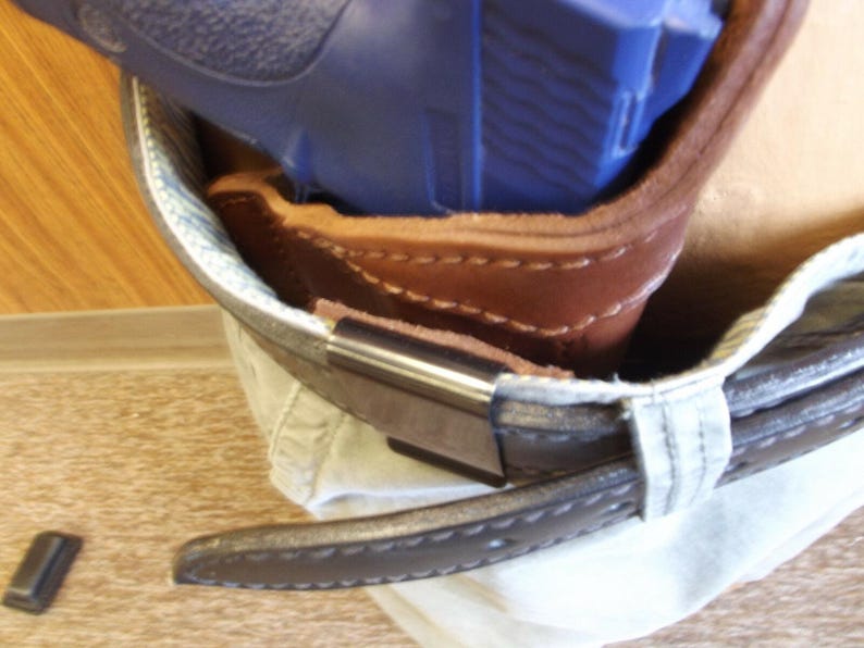 May include: A brown leather holster with a blue handgun inside, attached to a belt. The holster is stitched with visible seams and has a metal clip. The belt is dark brown with a stitched edge.