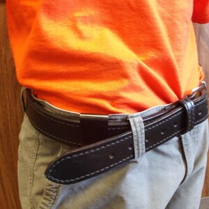 May include: A black leather belt with white stitching is fastened around the waist. The belt is worn over light-coloured trousers and under an orange top. The belt has a silver-coloured buckle.