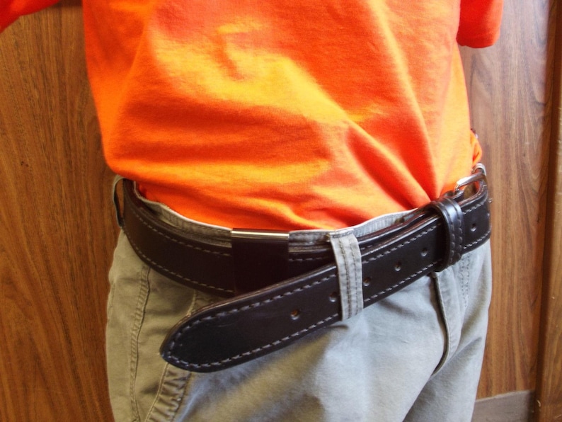 May include: A black leather belt with white stitching is fastened around the waist. The belt is worn over light-coloured trousers and under an orange top. The belt has a silver-coloured buckle.