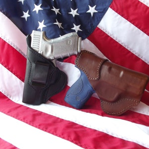 May include: Two leather holsters for handguns, one black and one brown, are displayed on a red, white, and blue American flag.
