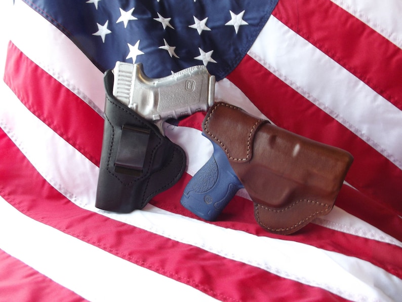 May include: Two leather holsters for handguns, one black and one brown, are displayed on a red, white, and blue American flag.