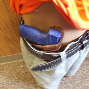 May include: A blue handgun is holstered in a brown leather holster, attached to a black belt. The belt is worn with light-coloured trousers. An orange shirt is visible above the belt.