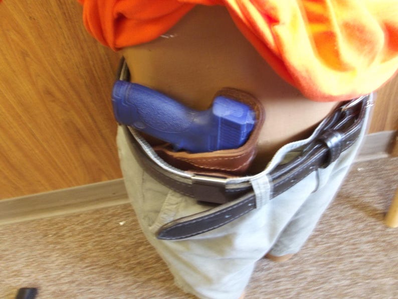 May include: A blue handgun is holstered in a brown leather holster, attached to a black belt. The belt is worn with light-coloured trousers. An orange shirt is visible above the belt.