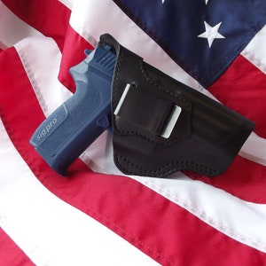 May include: A black leather holster with a silver clip, holding a blue and black handgun with the text "sig pro" on the side. The holster is lying on a red, white, and blue American flag.