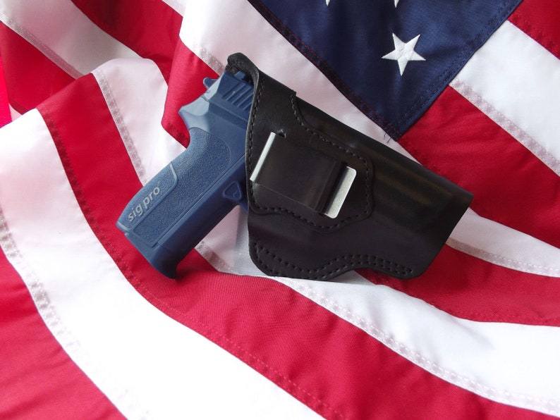 May include: A black leather holster with a silver clip, holding a blue and black handgun with the text "sig pro" on the side. The holster is lying on a red, white, and blue American flag.