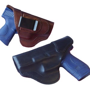 May include: Two leather gun holsters. One is brown with a black clip, and the other is black. Both holsters have a blue gun inside. The holsters are designed to securely hold a firearm.