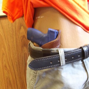 May include: A blue handgun in a brown leather holster, attached to a black leather belt with a silver buckle. The belt is worn with khaki trousers. An orange shirt is partially visible.
