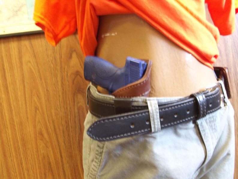 May include: A blue handgun in a brown leather holster, attached to a black leather belt with a silver buckle. The belt is worn with khaki trousers. An orange shirt is partially visible.