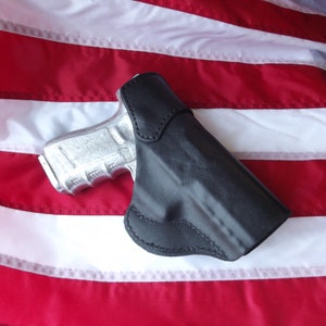 May include: A black leather holster for a handgun, with a silver handgun inside. The holster is lying on a red, white, and blue American flag.