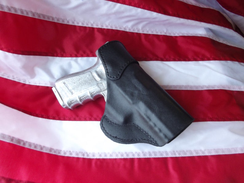 May include: A black leather holster for a handgun, with a silver handgun inside. The holster is lying on a red, white, and blue American flag.
