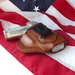 May include: A brown leather holster with a silver handgun inside. The holster is lying on a red, white, and blue American flag.