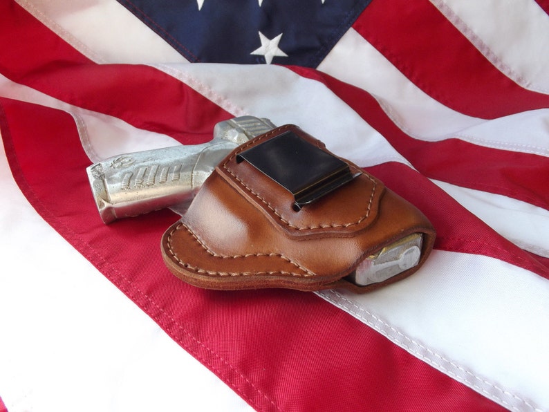 May include: A brown leather holster with a silver handgun inside. The holster is lying on a red, white, and blue American flag.