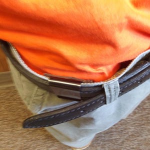 May include: Close-up of a brown leather belt with a silver buckle, worn over khaki trousers. An orange top is tucked into the trousers. The belt has a stitched design.