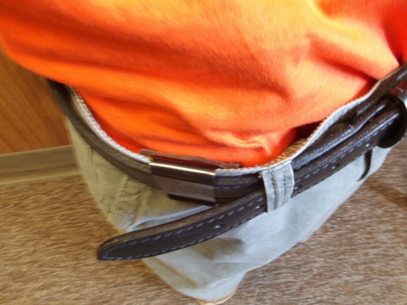 May include: Close-up of a brown leather belt with a silver buckle, worn over khaki trousers. An orange top is tucked into the trousers. The belt has a stitched design.