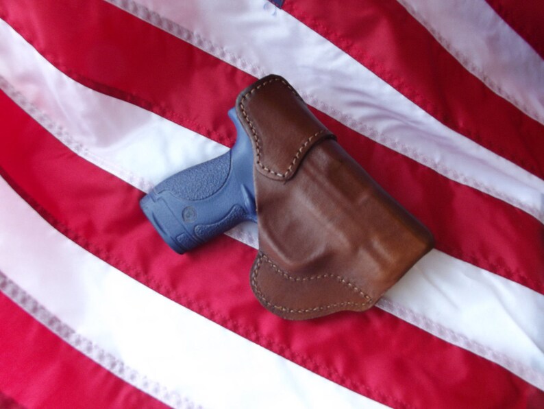 May include: A brown leather holster for a blue handgun, resting on a red, white, and blue American flag.