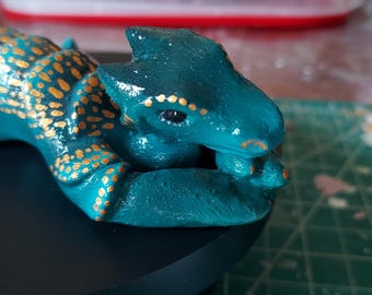 Op de afbeelding: Een keramiek drakenbeeldje, geschilderd in tealblauw en goud. De draak is opgerold met zijn kop rustend op zijn poten. De draak heeft gouden accenten op zijn lichaam en gezicht.