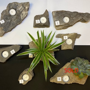 May include: Eight slate candle holders with a white candle in each. The candle holders are numbered 1 through 8. One candle holder has a succulent plant in it and is marked "SOLD".