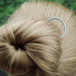 May include: A silver hair comb with a simple, curved design. The comb is holding a bun of light brown hair.