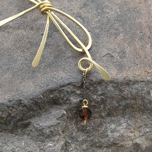 Op de afbeelding: Een gouden metalen strik met een hangende bruine kristallen hanger. De strik is gemaakt van draad en heeft een eenvoudig, elegant ontwerp.