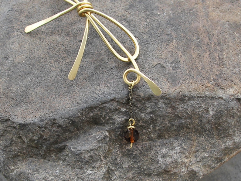 Op de afbeelding: Een gouden metalen strik met een hangende bruine kristallen hanger. De strik is gemaakt van draad en heeft een eenvoudig, elegant ontwerp.