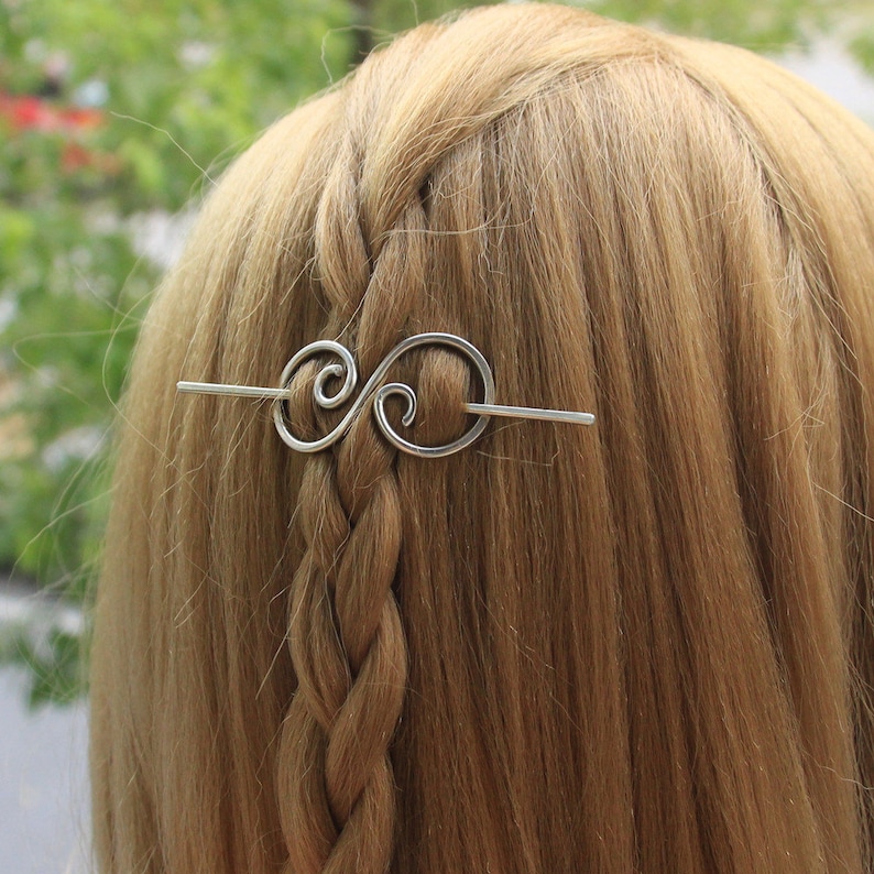 May include: Silver hair stick with a double swirl design. The stick is in a person's hair.