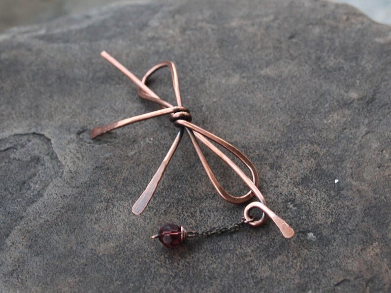 Op de afbeelding: Een haarspeld in de vorm van een strik van koperdraad met een hangende paarse glazen kraal en een koperen ketting.