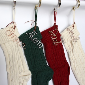 Puede incluir: Cuatro calcetines de Navidad de punto blanco, verde y rojo con nombres de alambre que dicen "Derek", "Papá", "Mamá" y "Abuela".