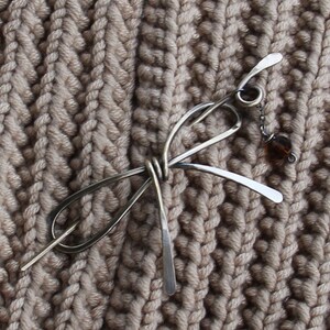 Op de afbeelding: Een zilveren haarspeld met een geknoopt ontwerp en een kleine bruine kraal die aan een ketting hangt. De speld ligt op een beige gebreide stof.
