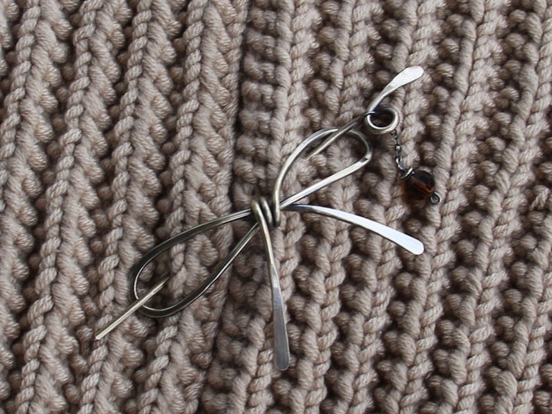 Op de afbeelding: Een zilveren haarspeld met een geknoopt ontwerp en een kleine bruine kraal die aan een ketting hangt. De speld ligt op een beige gebreide stof.