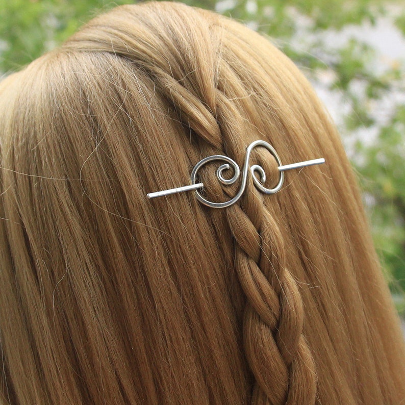 May include: A silver hair stick with a double swirl design. The stick is in a person's hair.