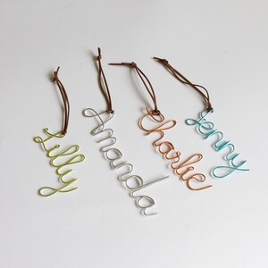 May include: Four wire name tags in different colors: green, silver, copper, and teal. Each tag features a different name: "Lilly", "Amanda", "Charlie", and "Jenny". The names are written in a cursive font and attached to brown leather cords.