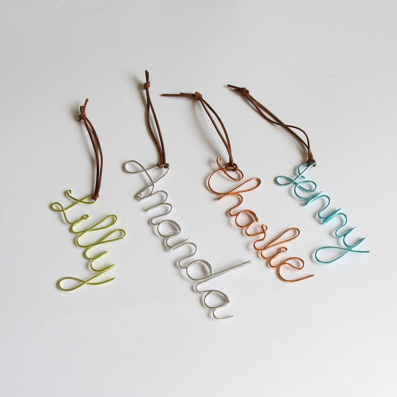 May include: Four wire name tags in different colors: green, silver, copper, and teal. Each tag features a different name: "Lilly", "Amanda", "Charlie", and "Jenny". The names are written in a cursive font and attached to brown leather cords.