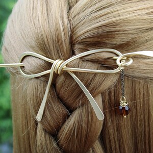 Op de afbeelding: Gouden metalen haarspeld in de vorm van een strik met een hangende bruine kraal aan een zwarte ketting.