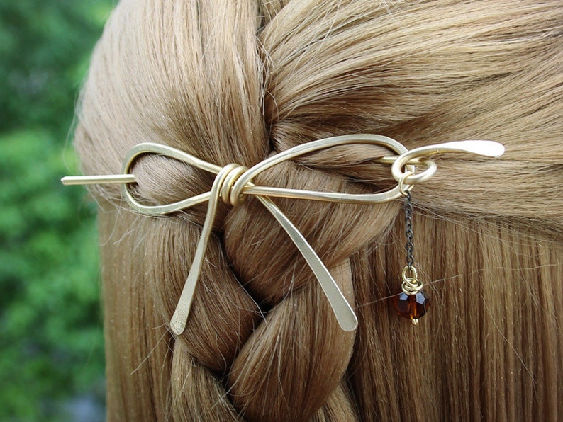 Op de afbeelding: Gouden metalen haarspeld in de vorm van een strik met een hangende bruine kraal aan een zwarte ketting.