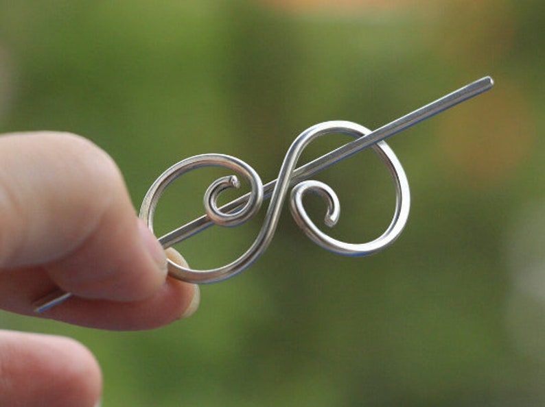 May include: A silver hair clip with a decorative swirl design. The clip is made of wire and has a simple, elegant look.