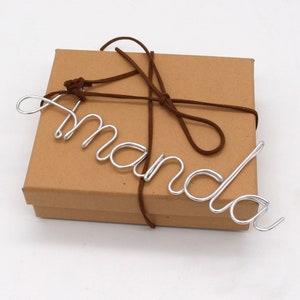 May include: A brown cardboard gift box tied with a brown suede cord. The name "Amanda" is crafted from silver wire and placed on top of the box. The box is on a white surface.