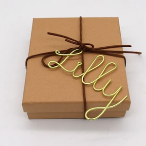 May include: A kraft paper gift box tied with a brown suede cord. The name "Lilly" is spelled out in gold wire and placed on top of the box. The box is square and has a simple, elegant design.
