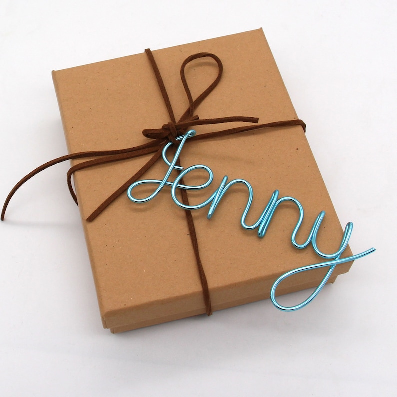May include: A brown cardboard box tied with a brown suede cord. A light blue wire name "Jenny" is placed on top of the box. The box is on a white surface.