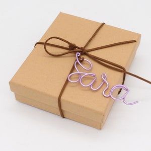 May include: A brown cardboard gift box tied with a brown suede cord. A lavender wire name tag that spells "Sara" is attached to the cord. The box is rectangular and has a simple, clean design.