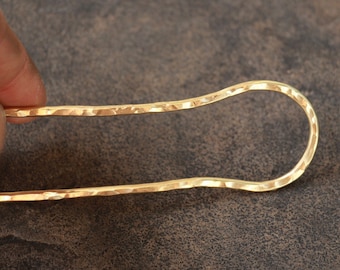 May include: A gold-colored metal hair stick with a hammered texture. The stick is curved and has a rounded end.