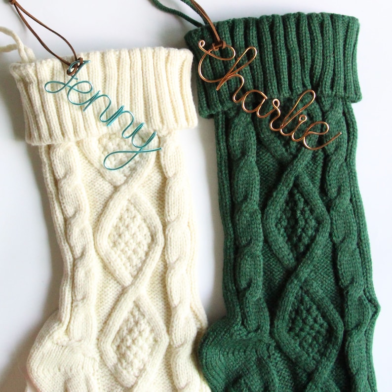 May include: Two cable-knit Christmas stockings, one cream and one dark green, with personalized wire name tags. The cream stocking has a teal wire name tag that says "Jenny." The green stocking has a copper wire name tag that says "Charlie."