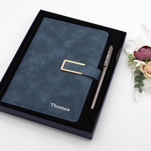May include: A blue faux-leather journal with a gold-coloured clasp and matching pen, presented in a dark blue box. The journal is embossed with the name "Thomas". A floral arrangement with a ribbon is on the right.