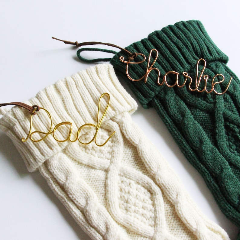 May include: Two cable-knit Christmas stockings, one cream and one dark green, with wire name tags that read "Dad" and "Charlie." The stockings have brown leather loops for hanging. The names are in gold and copper wire.