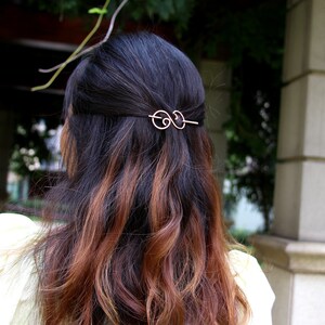 May include: A gold hair clip with a double loop design. The clip is in the hair of a person with long brown and black hair.